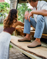 A man and a woman sitting on the back of a pickup truck outdoors. The man is wearing light blue jeans and taupe suede slip-on shoes, while the woman wears a brown top and light gray skirt.