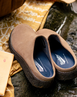 A pair of brown suede slip-on clogs with dark textured soles and blue insoles, styled on a wet dark stone surface with a yellow patterned fabric and a book in the background.