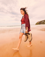 A woman walks barefoot on a sandy beach, holding a pair of taupe suede boots. She is wearing a red top, a dark burgundy button-up shirt, and denim shorts, with the ocean and cloudy sky in the background.