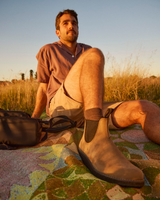 A man sitting outdoors in a grassy field at sunset, wearing tan Chelsea-style boots, beige shorts, and a mauve shirt. A patterned blanket is spread beneath him, with a dark bag beside him.