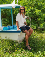 A man wearing a white short-sleeve button-up shirt, dark shorts, and sandals sits on the edge of a small white and blue boat in a grassy area with green foliage in the background.