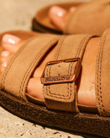 Close-up of a person wearing a pair of tan suede double-strap sandals with a brown buckle featuring the Blundstone logo, set against a textured concrete background.